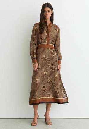 Kvinde stående iført en brun mønstret midi-kjole med lange ærmer, bælte i taljen og åbne hæle, stående mod en ensartet hvid væg.
