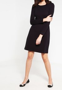 Svart långärmad klänning med rund halsringning och figurnära livstycke, som vidgar sig något vid fållen. Bärs med svarta lägenheter med metalliska detaljer.