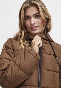 Brun polstret jakke med hætte, der har en frontzipper, sorte snore og quiltet struktur. Modellen holder om kraven.