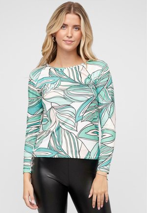 Haut à manches longues en blanc et vert menthe, affichant un motif de feuilles et un col arrondi ; porté avec des leggings noirs. Texture de tissu lisse.