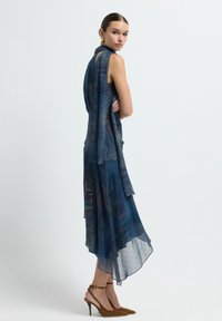 Vestido fluido de color azul oscuro con tela transparente, dobladillo asimétrico y patrones intrincados. Combinado con zapatos de tacón alto de ante marrón.