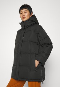 Svart dunjakke med høy krage, glidelås foran og sidelommer. Den teksturerte stoffet har quiltede seksjoner og en avslappet passform.