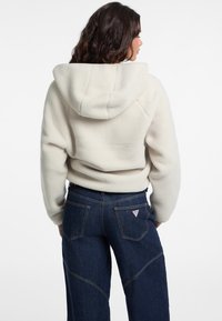 Giacca a cappuccio bianca in fleece con una texture morbida, abbinata a jeans a vita alta blu scuro con un logo triangolare rosso sulla tasca posteriore.