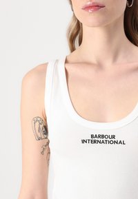 Valge ribadega tank top, millel on ümar kaelus ja musta tikandiga "BARBOUR INTERNATIONAL" tekst ees. Sile kangastekstuurne.