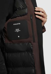 Melns un brūns pūkains jaku ar rāvējslēdzēja kabatu, kurā ir logo teksts, gluda melna auduma virsma un quiltēts siltinājums.