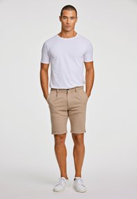 Biała koszulka z krótkim rękawem w zestawieniu z beżowymi, podwiniętymi szortami oraz białymi tenisówkami. Model stoi prosto, z rękami w kieszeniach, na jasnym tle.