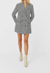 Vestito blazer doppiopetto a motivo houndstooth bianco e nero con rever e quattro bottoni, abbinato a stivaletti beige alla caviglia.