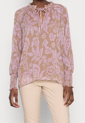 Femme portant un chemisier beige à manches longues avec un motif floral rose, des poignets froncés et un détail noué au col, associé à un pantalon beige clair.
