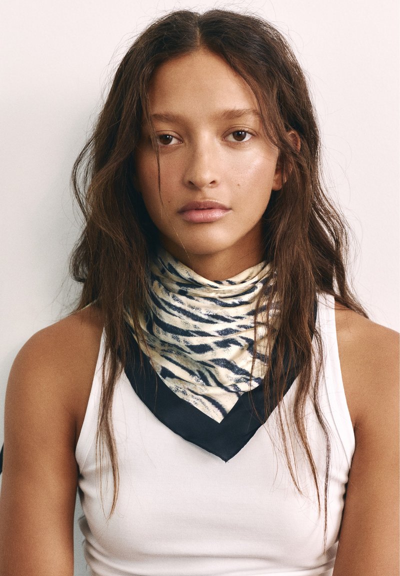 Jeune femme aux longs cheveux bruns portant un débardeur blanc et une écharpe à motif zèbre autour du cou, face à l’avant avec une expression neutre.