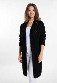 Cardigan negru cu tricotaj din fire groase, lung, cu o croială lejeră, model texturat, deschis în față și cu mâneci până la trei sferturi, purtat peste un top alb și pantaloni.