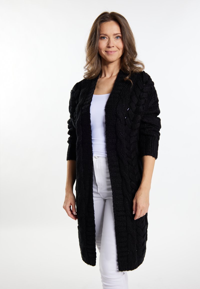 Cardigan negru cu tricotaj din fire groase, lung, cu o croială lejeră, model texturat, deschis în față și cu mâneci până la trei sferturi, purtat peste un top alb și pantaloni.