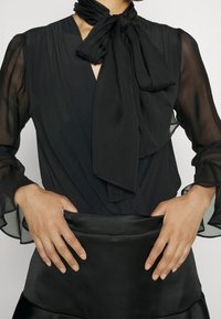 Black blouse with sheer sleeves and a tied neck scarf, paired with a smooth, fitted satin skirt. Both garments feature a fluid texture.