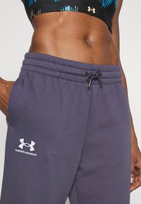 Mørkegrå sweatpants med elastisk talje og snørre. Har sidelommer og hvidt broderet "Under Armour"-logo. Glat tekstur.