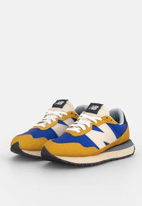 Paire de baskets New Balance en bleu, jaune moutarde, blanc et noir avec des lacets blancs et un logo sur la languette et le côté.