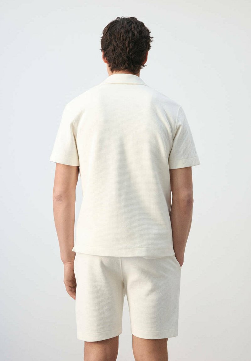 Hombre de pie de espaldas con camisa blanca texturizada de manga corta y pantalones cortos a juego sobre un fondo claro liso.