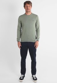 Chemise verte à manches longues avec un col rond et un petit logo. Associée à un pantalon bleu marine et des chaussures noires avec des semelles blanches. Texture lisse.