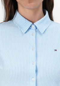 Camicia a righe azzurro chiaro con colletto classico, chiusura frontale con bottoni e piccolo logo sul lato sinistro. Realizzata in un tessuto texturizzato.