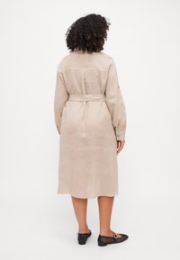 Abito in lino beige con vita a fascia, maniche lunghe e chiusura con bottoni sul retro. Indossato con ballerine in rete nere con un accento circolare.
