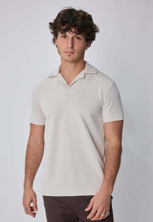 Jeune homme aux cheveux bouclés portant une chemise beige à manches courtes avec col et un pantalon foncé, debout devant un fond clair uni.