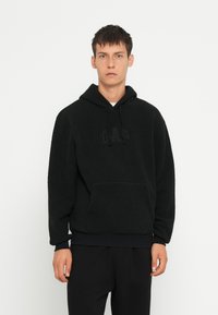 Felpa nera in pile con tasca a marsupio, caratterizzata da un discreto logo "GAP" impresso. Ha un cappuccio regolabile con lacci e polsini a coste.