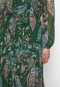 Vestido verde con patrón de paisley en azul, blanco y naranja, mangas largas transparentes, lazo en la cintura y detalle de falda fruncida, mano descansando al lado.