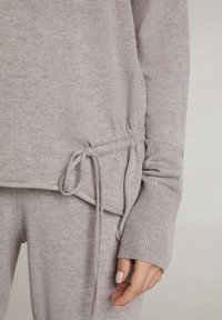 Detailní záběr na ruku osoby ležící vedle těla, oblečenou v světle šedém pleteném svetr a kalhoty s šňůrkou uvázanou v pase.