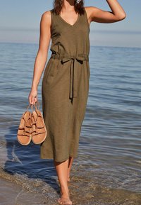 Robe maxi sans manches de couleur vert olive avec un décolleté en V et une taille à cordon, associée à des sandales à lanières beige tenues dans une main, sur une plage de sable.