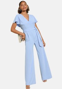 Combinaison légère bleue avec des manches courtes en volants, décolleté en V profond, taille ceinturée et pantalons à jambes larges ; tissu lisse avec un design épuré.