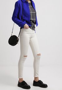 Giacca corta blu, top grigio con grafica, jeans skinny bianchi strappati e scarpe nere con lacci. Borsa a tracolla rotonda nera con accessori dorati.