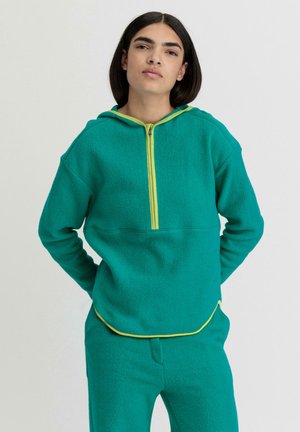 Türkisfarbener Fleece-Hoodie mit gelbem Reißverschluss und Besatz, ausgestattet mit einer Fronttasche, lockerer Passform und Kapuze. Kombiniert mit türkisfarbenen Fleece-Hosen.