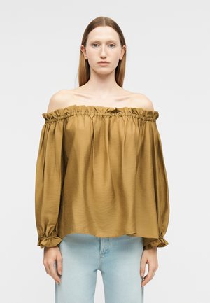 Egyenes barna hajú nő laza mustársárga vállra húzott blúzt és világoskék farmert visel, tiszta világos háttér előtt áll.