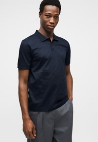 Μπλε ναυτικό πόλο με υφασμάτινο σχέδιο, κοντά μανίκια, τρεις κουμπότρυπες και άνετη εφαρμογή. Φορεμένο με ραμμένο γκρι παντελόνι.