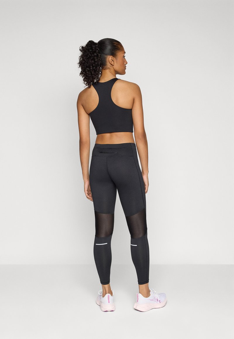 Schwarzer Sport-BH mit Racerback-Design, kombiniert mit hochgeschnittenen schwarzen Leggings, die Netzeinsätze und reflektierende Akzente aufweisen. Leichtes Schuhwerk.