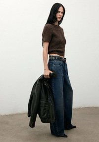 Suéter marrón corto con mangas cortas, jeans de mezclilla azul de cintura alta, chaqueta de cuero negra colgada del brazo y zapatos de tacón negros.