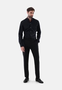 Schwarzes Hemd mit schmaler Passform und Mandarin-Kragen, kombiniert mit schmal geschnittenen schwarzen Hosen und passenden schwarzen Schuhen, vor einem neutralen Hintergrund.