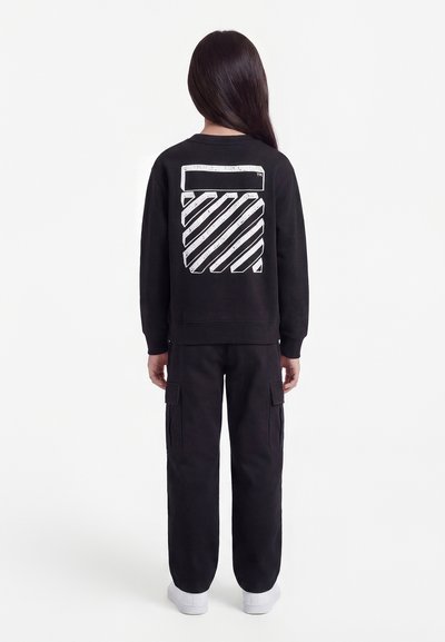 Παιδί που κοιτάζει μακριά, φοράει μαύρο φούτερ με λευκό γραφικό διαγώνιας ρίγας στην πλάτη, μαύρο παντελόνι cargo και λευκά αθλητικά παπούτσια.