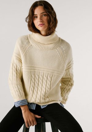 Jersey de punto grueso de color crema con cuello de tortuga, que presenta patrones de punto de cable y acentos acanalados. Lleva puesto sobre una camisa a rayas azules.