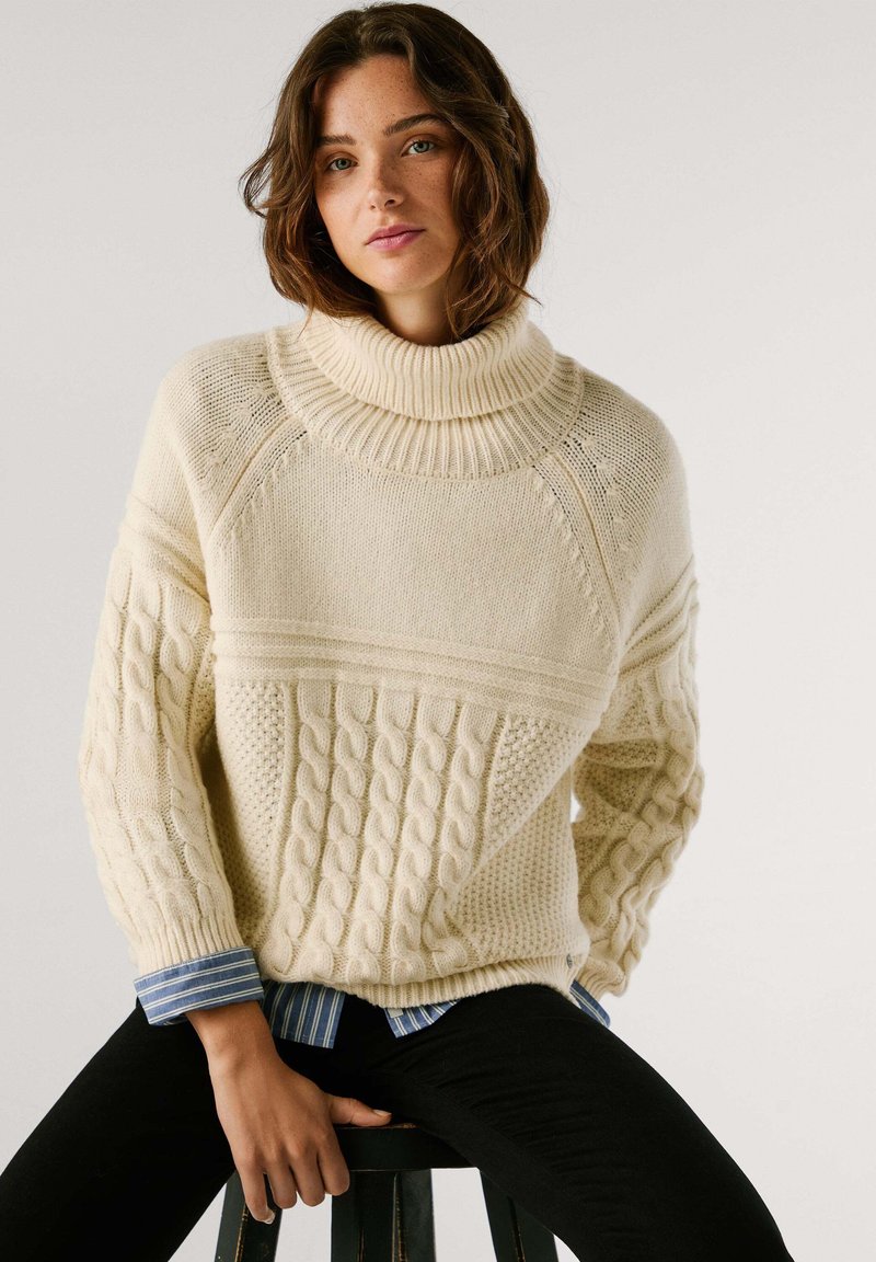 Chunkig krämfärgad stickad tröja med hög krage, som har kabelstickade mönster och ribbade detaljer. Bärs över en blårandig skjorta.