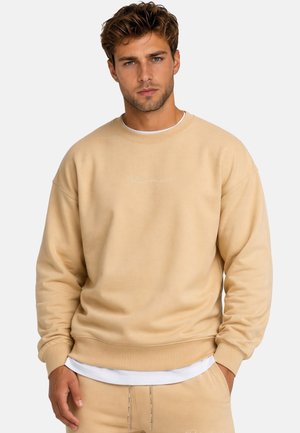Giovane uomo che indossa una felpa beige e pantaloni coordinati con una mano in tasca, in piedi davanti a uno sfondo chiaro e uniforme.