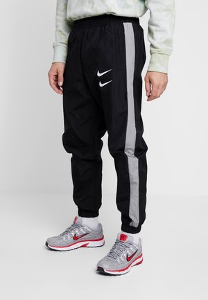 Personne portant un pantalon de survêtement Nike noir avec des bandes latérales grises et des baskets Nike grises avec des accents rouges, debout sur un fond uni.