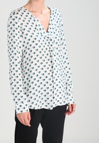 Long-sleeve blouse in lightweight, sheer fabric featuring a white background with teal heart patterns and a V-neck design.