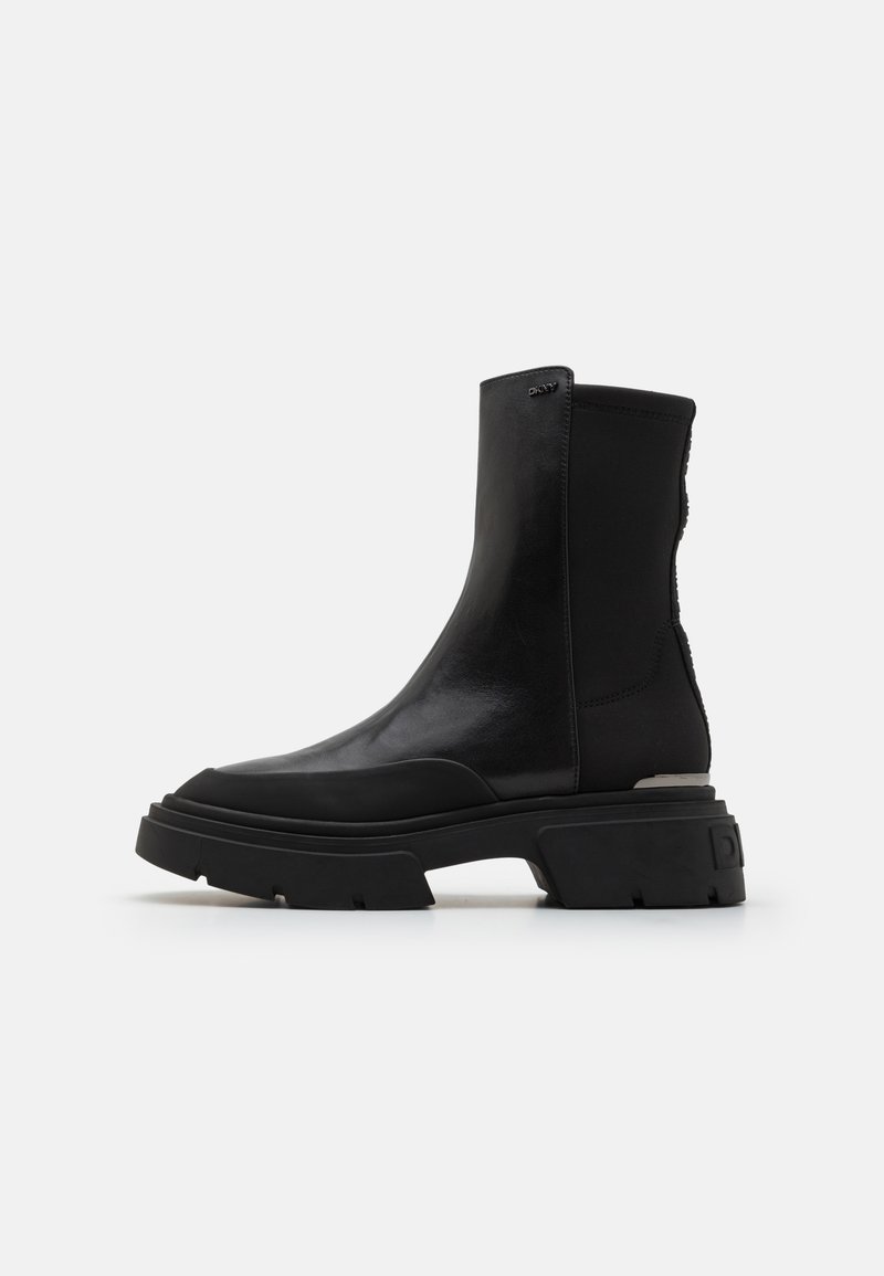 Botas negras hasta el tobillo con un superior de cuero brillante, paneles elásticos en los laterales, suela de goma gruesa y acentos texturizados en contraste.