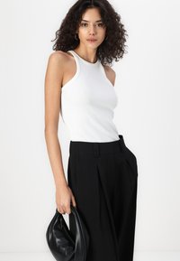White ribbed tank top paired with high-waisted black trousers, featuring a pleated design and a smooth leather bag in hand.