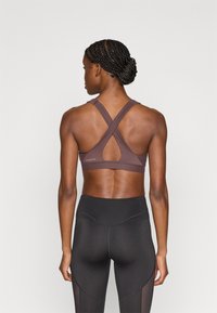 Καφέ αθλητικό σουτιέν με λουριά που διασταυρώνονται στην πλάτη, με λεία υφή και ελάχιστη σήμανση. Συνδυάζεται με μαύρο κολάν ψηλής μέσης.