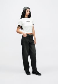 Νεαρή γυναίκα που φοράει λευκό crop top, μαύρο παντελόνι με φαρδιά μπατζάκια, μαύρα αθλητικά παπούτσια και γκρι καπέλο με το γείσο πίσω, όρθια με το ένα χέρι στην τσέπη.