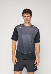 Young man with curly hair stands facing forward wearing a black and gray sports jersey and black athletic shorts with Nike logos.