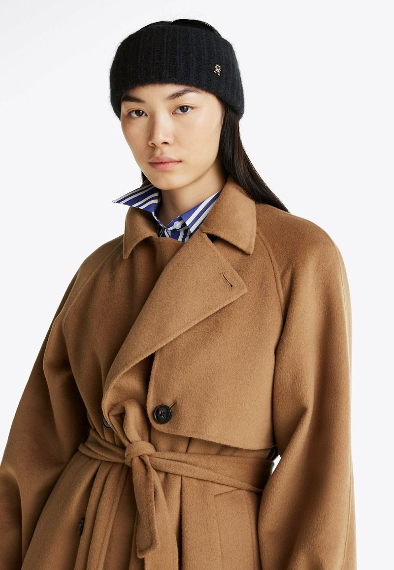 Manteau en mélange de laine marron avec un design enroulé, deux grands boutons noirs et une ceinture à nouer. En dessous, une chemise à col rayée bleu et blanc.