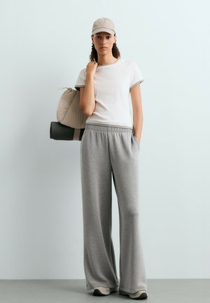 Jeune femme avec une casquette beige, un t-shirt blanc et un pantalon large gris, tenant un tapis beige roulé et un tapis de yoga noir contre un mur clair.