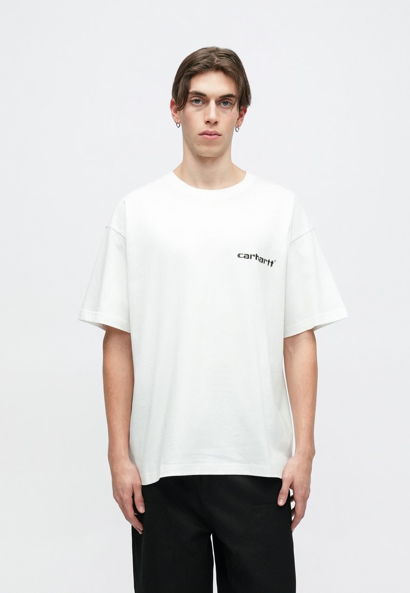 Camiseta de algodón blanca con un corte relajado, mangas cortas y un pequeño logo negro de "carhartt" en el lado izquierdo. Combinada con pantalones negros.