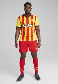 Puma fodboldudstyr med en gul og rød stribet trøje med sorte detaljer, røde shorts og matchende sokker med logo.